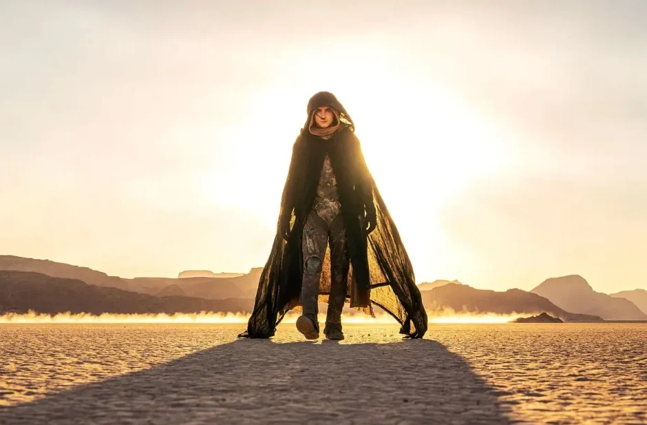 Timothée Chalamet as Paul Atreides walking forward across a sun-drenched desert expanse, wearing a dark hooded cloak and stillsuit with a bright, hazy sky behind him.