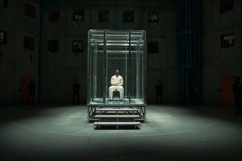 Assane Diop seated inside a transparent glass enclosure in the center of a high-security prison facility in Lupin Part 4.