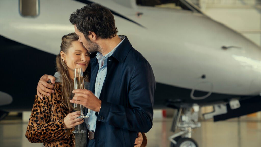 Kristen Bell as Joanne and Adam Brody as Noah embracing and holding champagne flutes in front of a private airplane in a scene from Nobody Wants This.