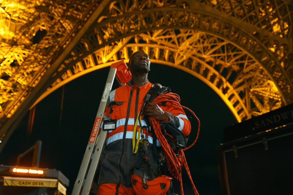Omar Sy as Assane Diop wearing an orange high-visibility jumpsuit and carrying climbing gear beneath the illuminated Eiffel Tower at night in Lupin Part 4.